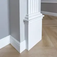 Biały pilaster z żłobieniami, ozdobny cokół i listwy przypodłogowe na tle szarej ściany i drewnianej podłogi w jodełkę.