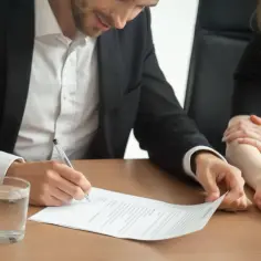 Mężczyzna w garniturze podpisuje dokument, który może być pełnomocnictwem ogólnym. Obok stoi kobieta.