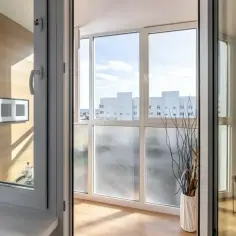 Zabudowa balkonu w bloku z widokiem na białe budynki. Wnętrze z drewnianą ścianą i wazonem z gałązkami.