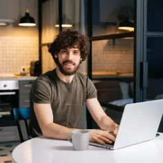 Uśmiechnięty mężczyzna z kręconymi włosami pracuje na laptopie przy stole w kuchni. Może właśnie sprawdza, czy wystarczy mu na czynsz.