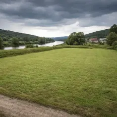 Łąka nad rzeką, domy w oddali. Chmury zapowiadają deszcz, który może spowodować **powódź**.