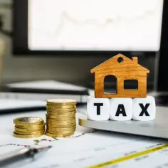 Drewniany domek na kostkach z napisem TAX, obok stosy monet. Symbolizuje podatek od czynności cywilnoprawnych przy zakupie nieruchomości.
