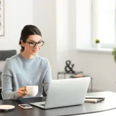 Kobieta w okularach pije kawę, pracując na laptopie przy biurku.