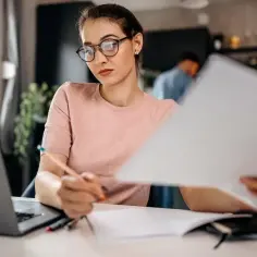 Kobieta w okularach pracuje nad dokumentami, przygotowując wniosek o wykreślenie hipoteki. Obok laptop i notatnik.