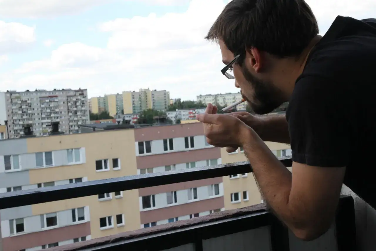 Mężczyzna z papierosem w ustach, zapalający go na balkonie. W tle blokowisko. Czy można palić na balkonie?