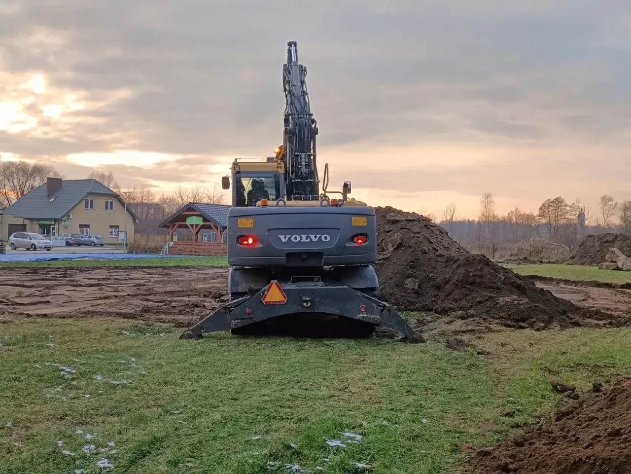 Koparka Volvo pracuje przy niwelacji terenu. W tle budynki i zachmurzone niebo.