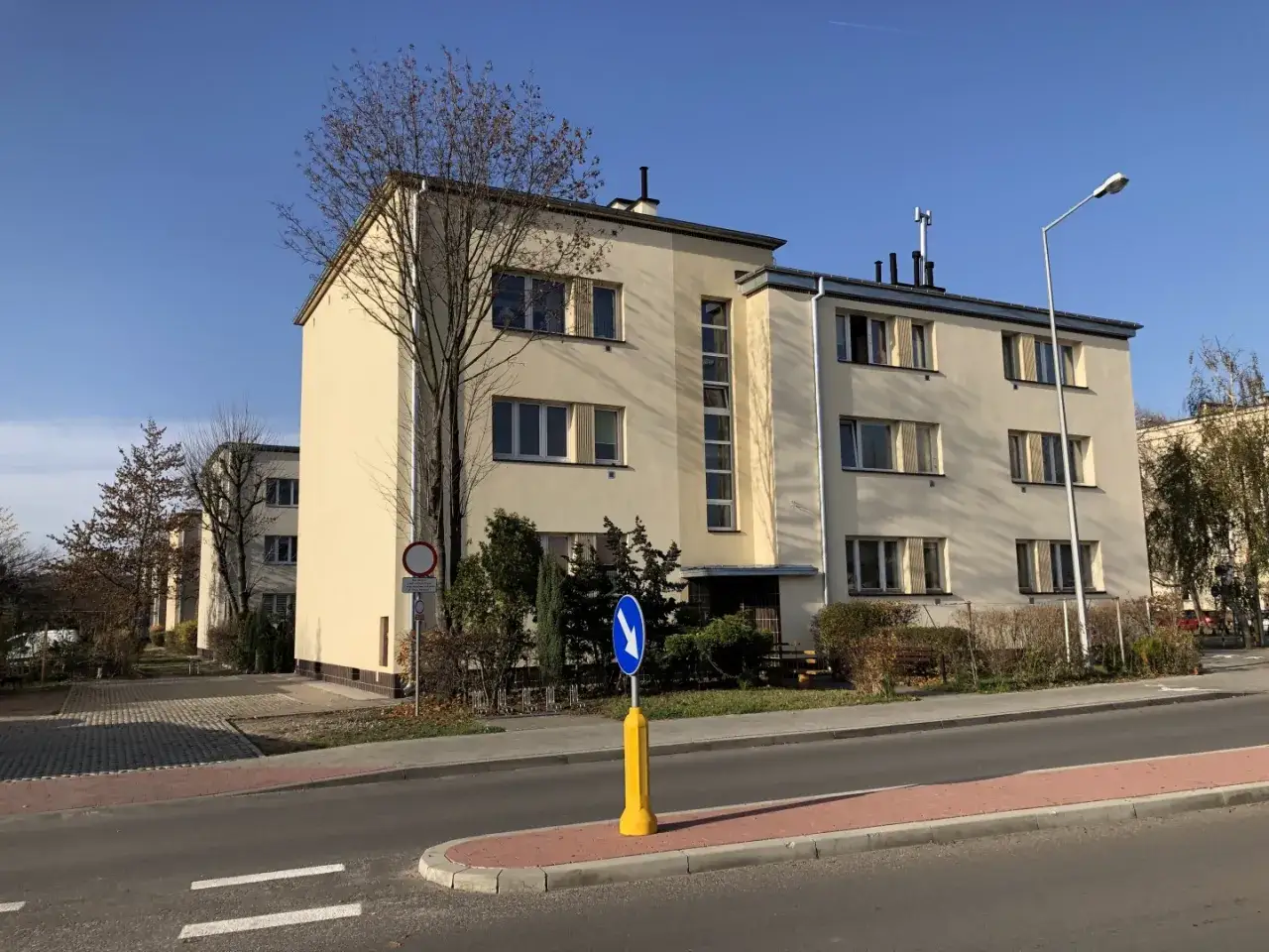 Jasny budynek mieszkalny w stylu art deco z geometrycznymi oknami i prostymi liniami, otoczony drzewami.