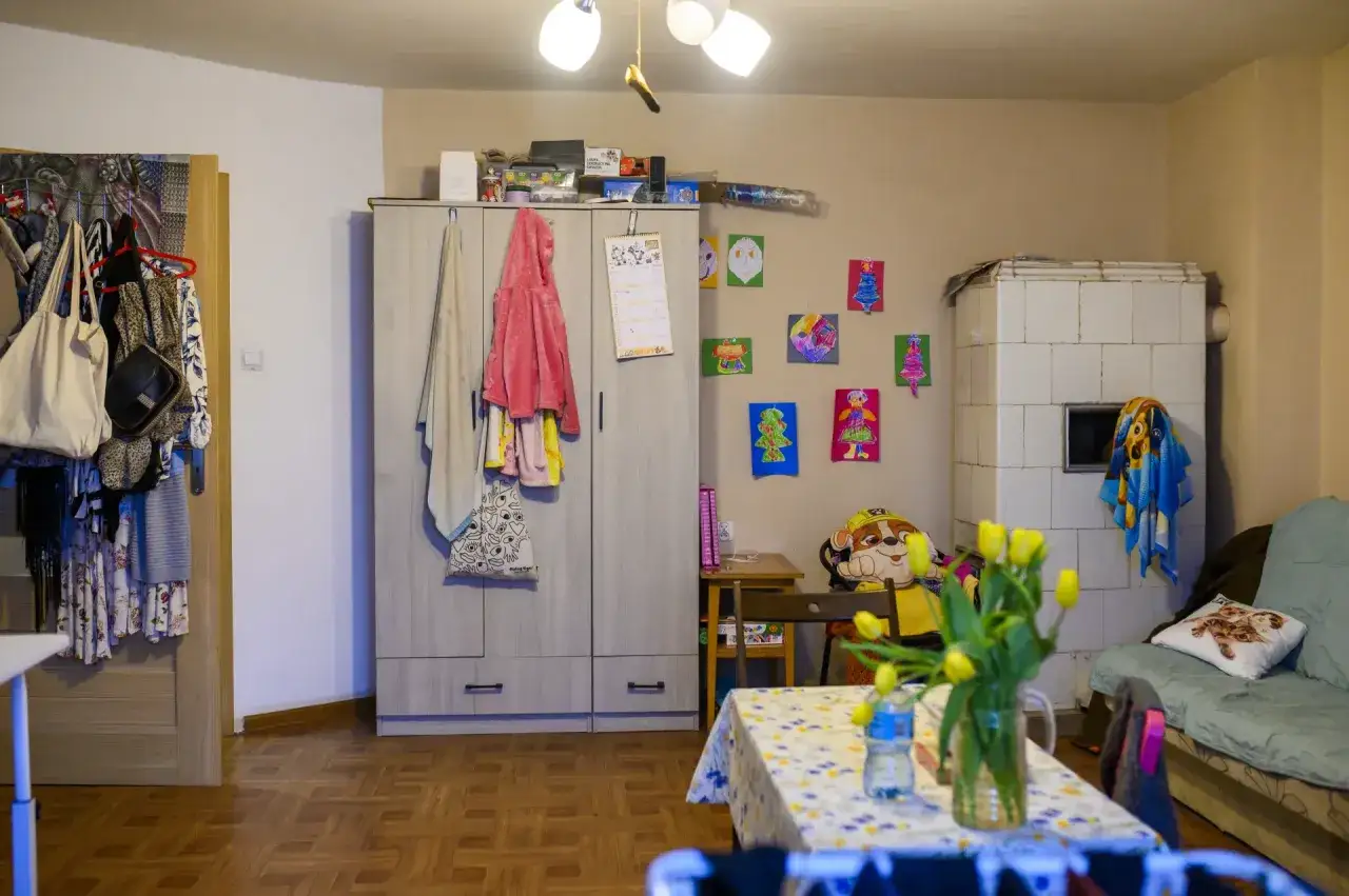 Przytulne mieszkanie socjalne: szafa z ubraniami, dziecięce rysunki na ścianie, piec kaflowy i kanapa.